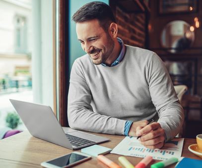 Man in grijze trui, lachend en aan het werk in café, kijkend naar laptop