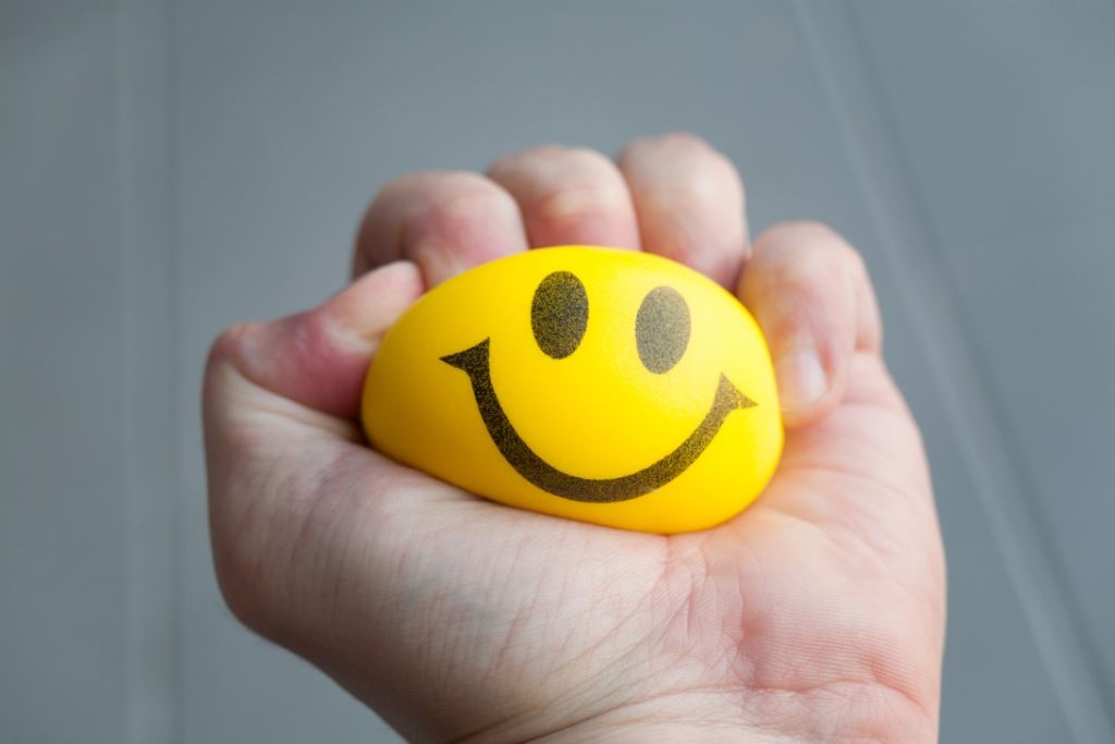 Een hand die een gele stressbal met een smiley erop fijnknijpt.