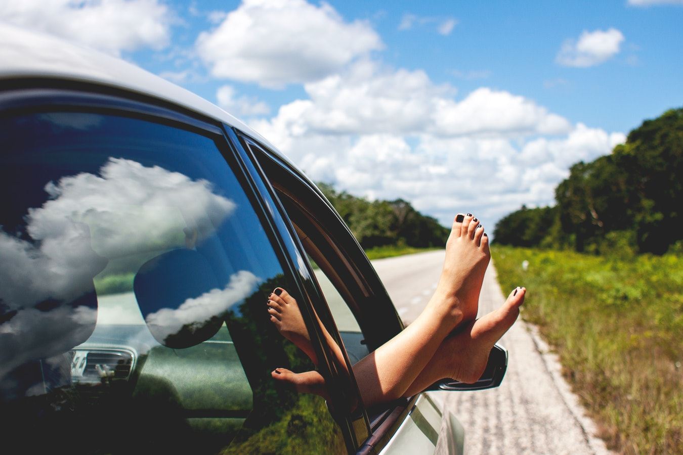 Een vrouw steek haar voeten uit de auto. Naast de weg waar de auto op stilstaat is een berm die overloopt in een bos.