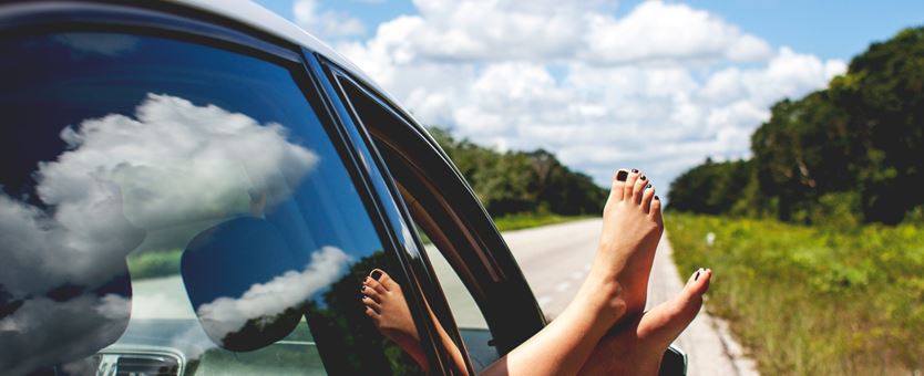 Een vrouw steek haar voeten uit de auto. Naast de weg waar de auto op stilstaat is een berm die overloopt in een bos.