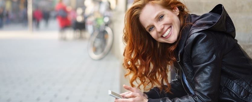 Een vrouw met rode haren zit buiten op een trapen kijkt lachend de camera in, in haar hand heeft ze haar telefoon.