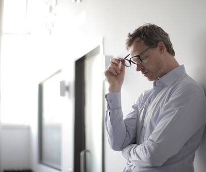 Een man staat te leunen tegen een muur en houd zijn zwarte bril vast.