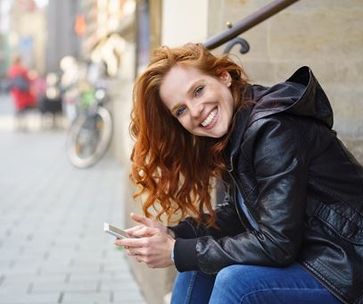 Een vrouw met rode haren zit op een trap en glimlacht, ze heeft haar telefoon in haar hand