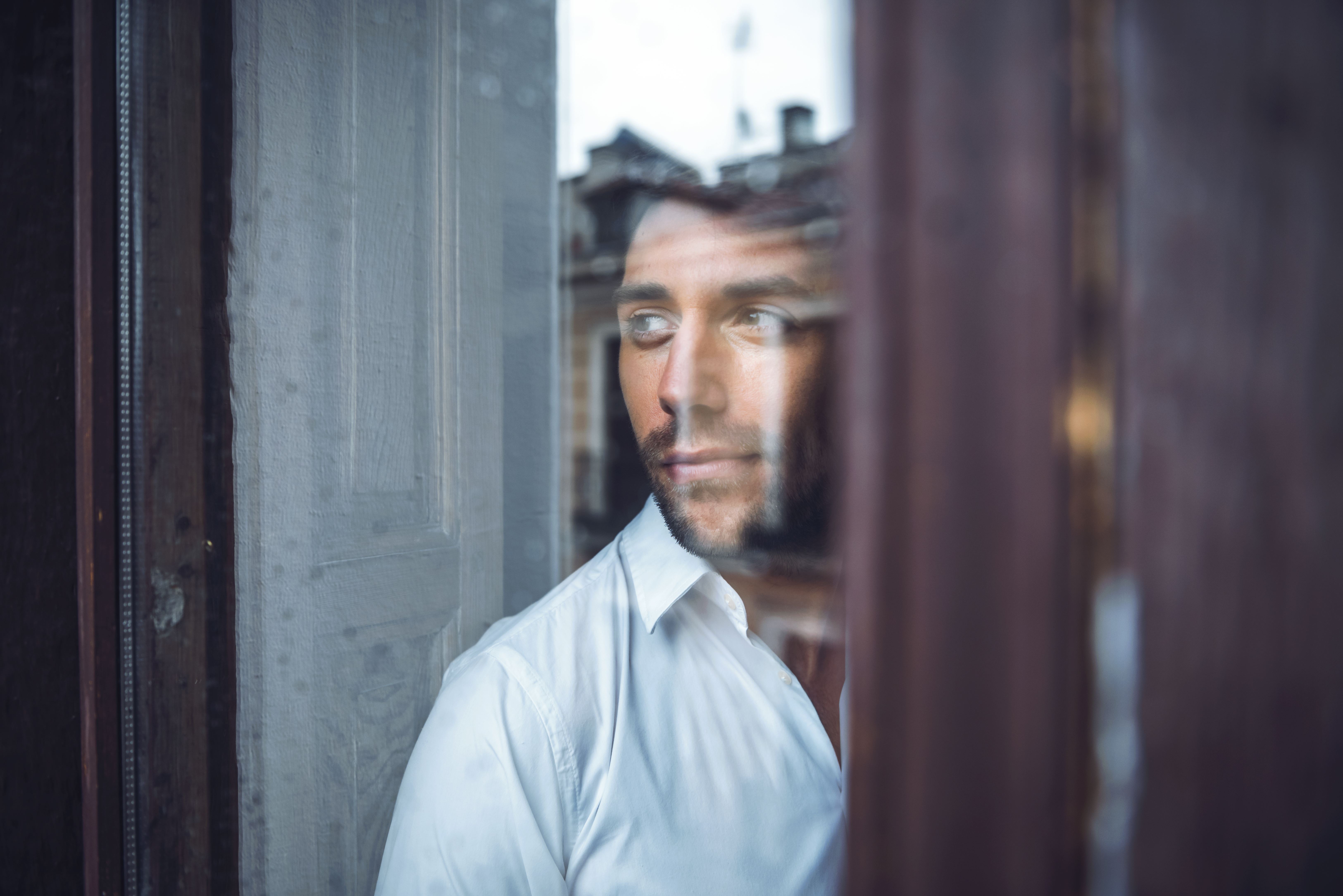 Een man met een witte blouse staat achter een raam en kijkt naar buiten.