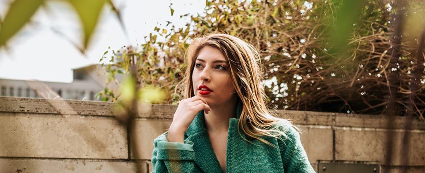 Een vrouw met een groene jas, rode lippenstift en bruine haren zit buiten en denkt na.