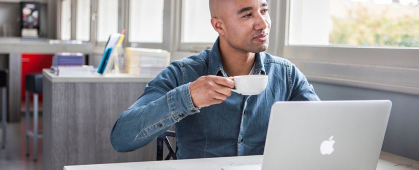 Een man werkt op zijn laptop en drinkt een kopje koffie.