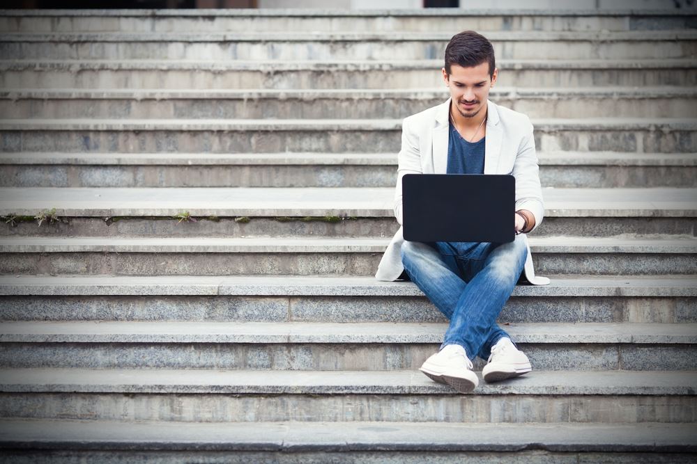 Een man zit buiten op de trap en werkt op zijn laptop, hij heeft een witte jas en een blauwe broek aan.