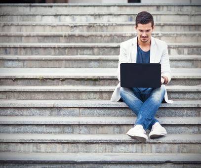 Een man zit buiten op de trap en werkt op zijn laptop, hij heeft een witte jas en een blauwe broek aan.