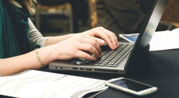 Een vrouw typt op haar laptop, naast haar ligt een papiertje en haar telefoon.