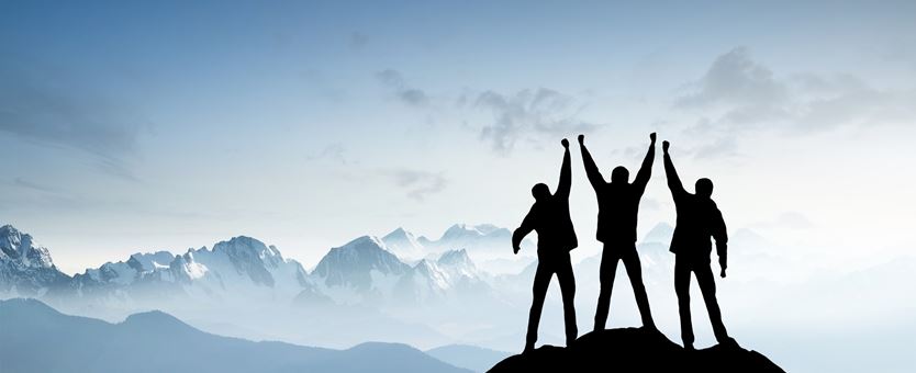 Drie mensen staan op een berg, de mensen hebben alle drieën handen in de lucht en juichen.
