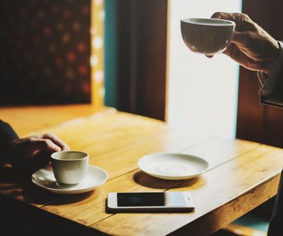 Twee mensen staan aan een houte tafel koffie te drinken. Op tafel staan de twee kopjes koffie en een telefoon.