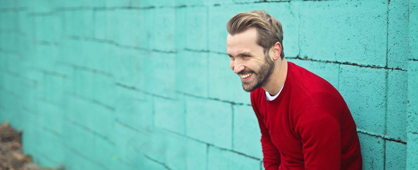 Een man met een rode trui staat lachend voor een turquoise stenen muur.