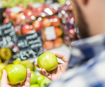 Een man staat in de supermarkt bij de appelafdeling. De man staat twee groene appels met elkaar te vergelijken.