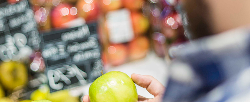 Een man staat in de supermarkt bij de appelafdeling. De man staat twee groene appels met elkaar te vergelijken.