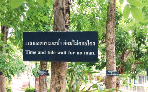 Een bord hangt aan de boom. Op het bord staat: Time and tide wait for no man. Daarboven staat dezelfde tekst in het Thais.