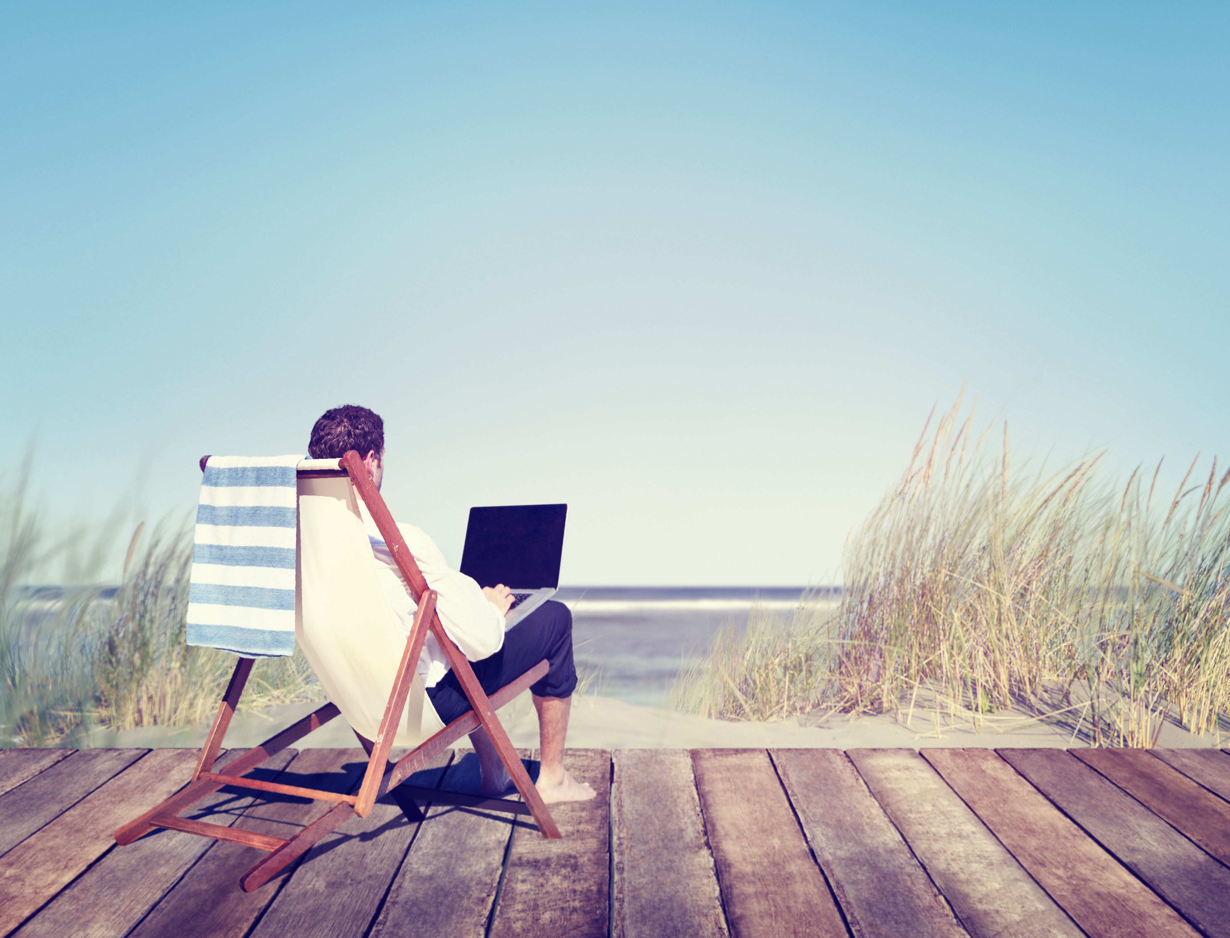 Een man zit aan het strand in een strandstoel te werken op zijn laptop.