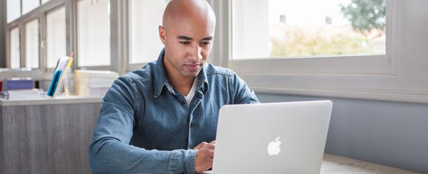 Een man zit aan een tafel, bij het raam en is aan het werk op zijn laptop. Naast de laptop staat een kopje koffie.