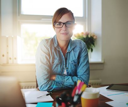 Een vrouw met oranje haren zit achter haar bureau en kijkt in de camera, op het bureau staat haar laptop en notitieboekjes.