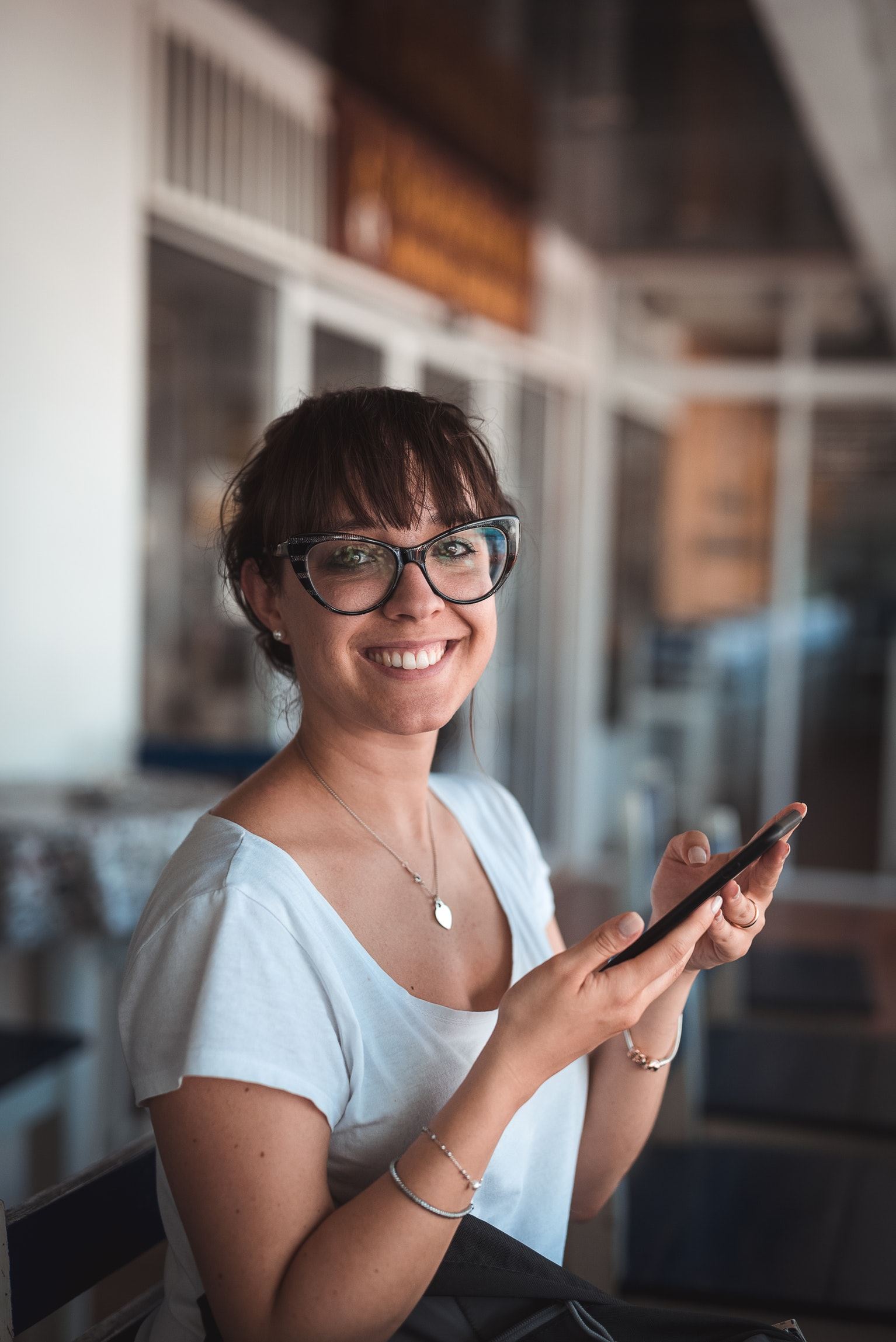 Vrouw met een grote bril heeft haar telefoon in haar hand en lacht in de camera.