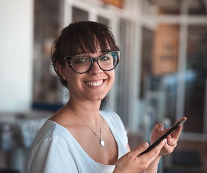 Vrouw met een grote bril heeft haar telefoon in haar hand en lacht in de camera.