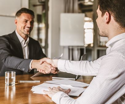 Twee mannen in pak tegenover elkaar aan een bureau schudden elkaar de hand voor een deal.