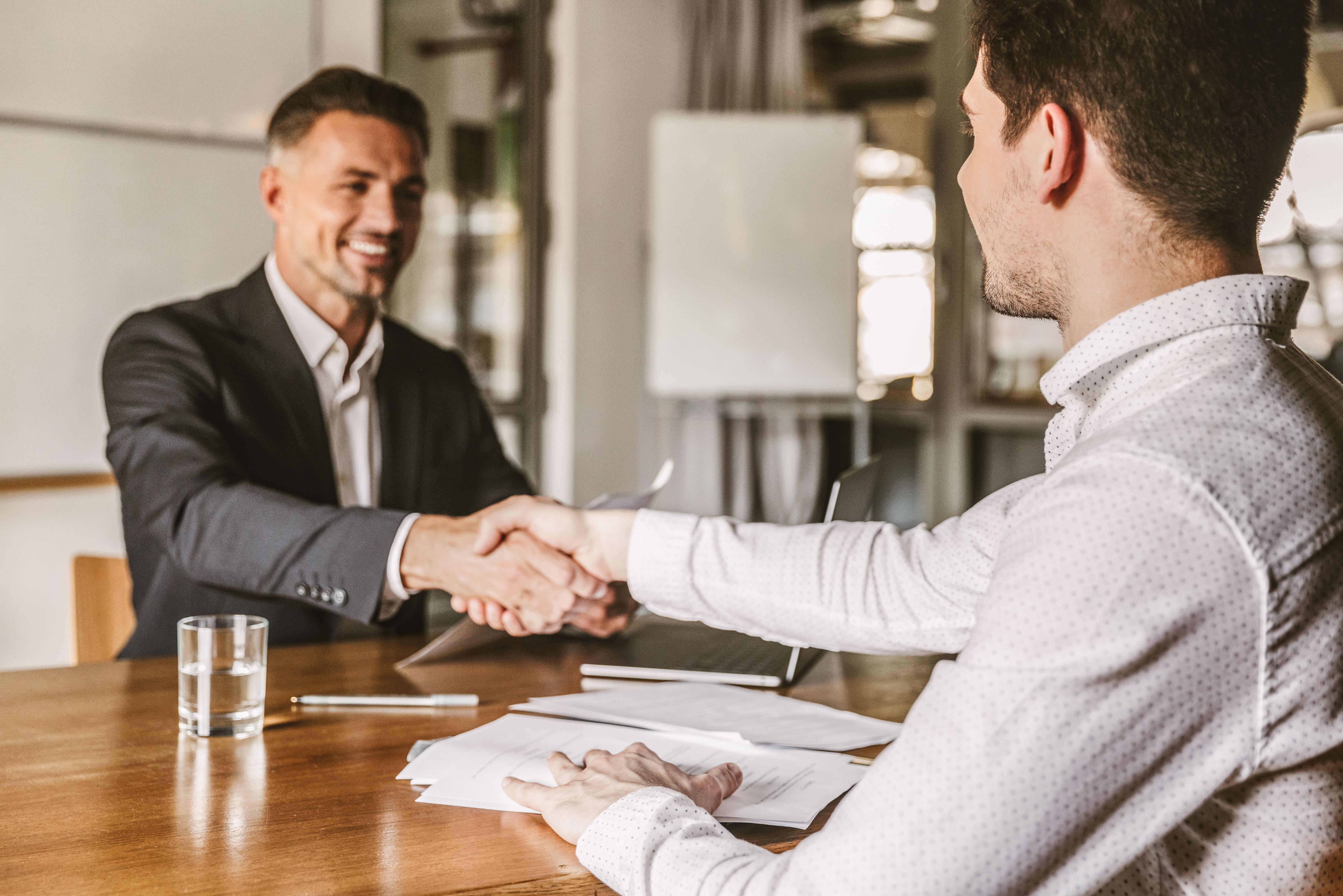 Twee mannen in pak tegenover elkaar aan een bureau schudden elkaar de hand voor een deal.