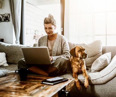Een vrouw zit in de kleermakerszit op de bank met haar laptop op schoot, naast haar op de bank ligt haar hond.
