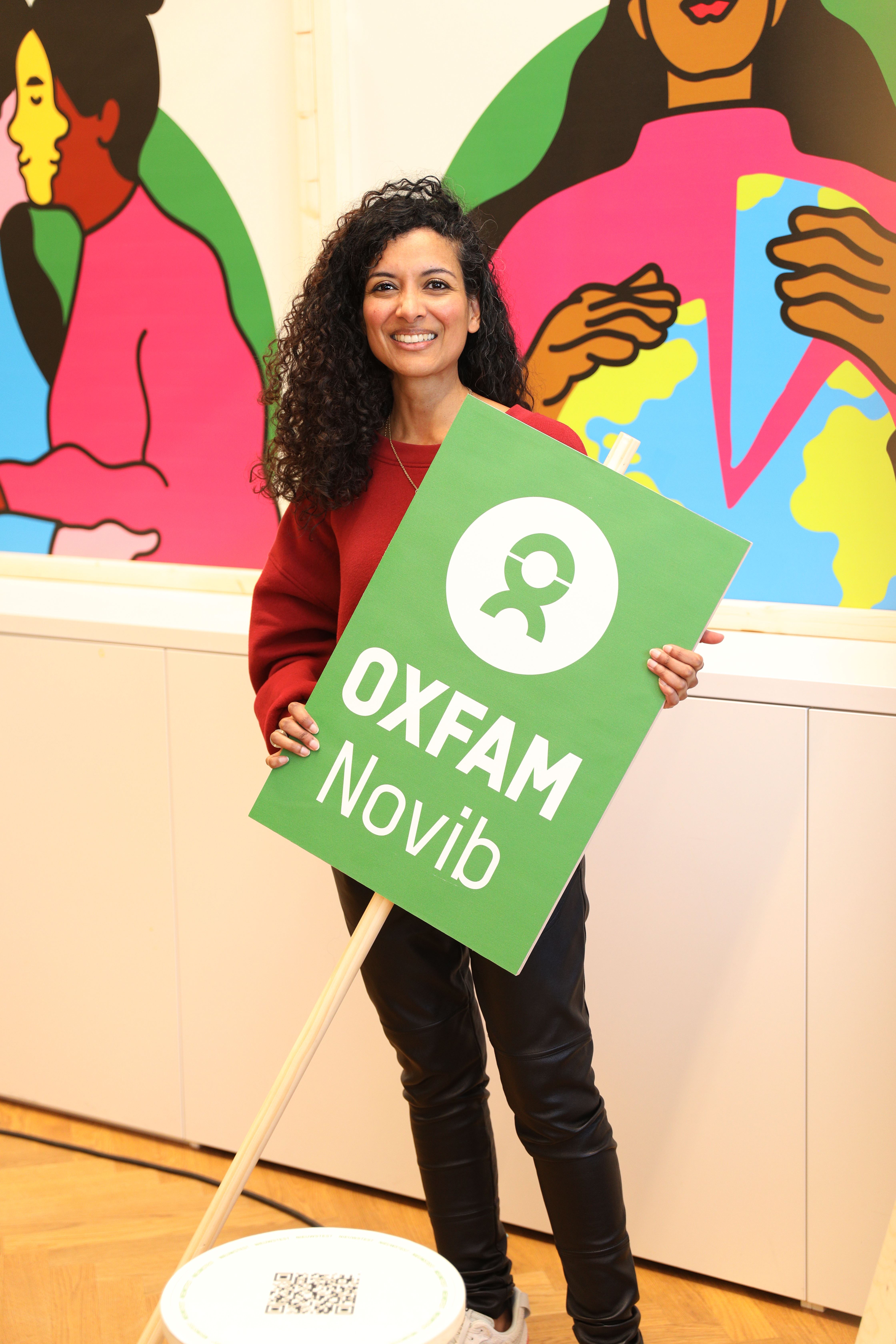 Een vrouw met donkere haren en een rode trui heeft een groen bord vast waar Oxfam Novib op staat.