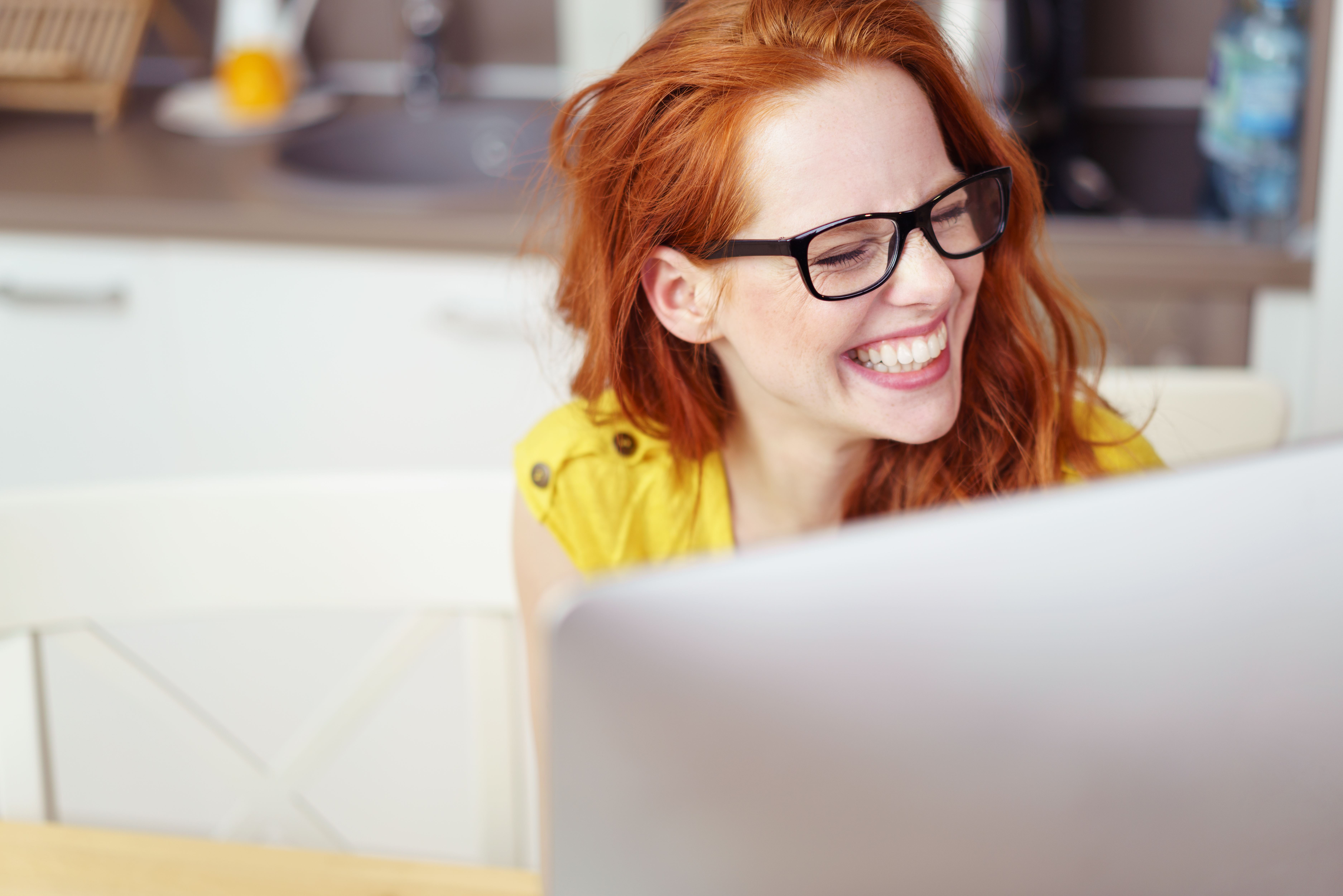 Een vrouw met rode haren zit achter haar computer en lacht.
