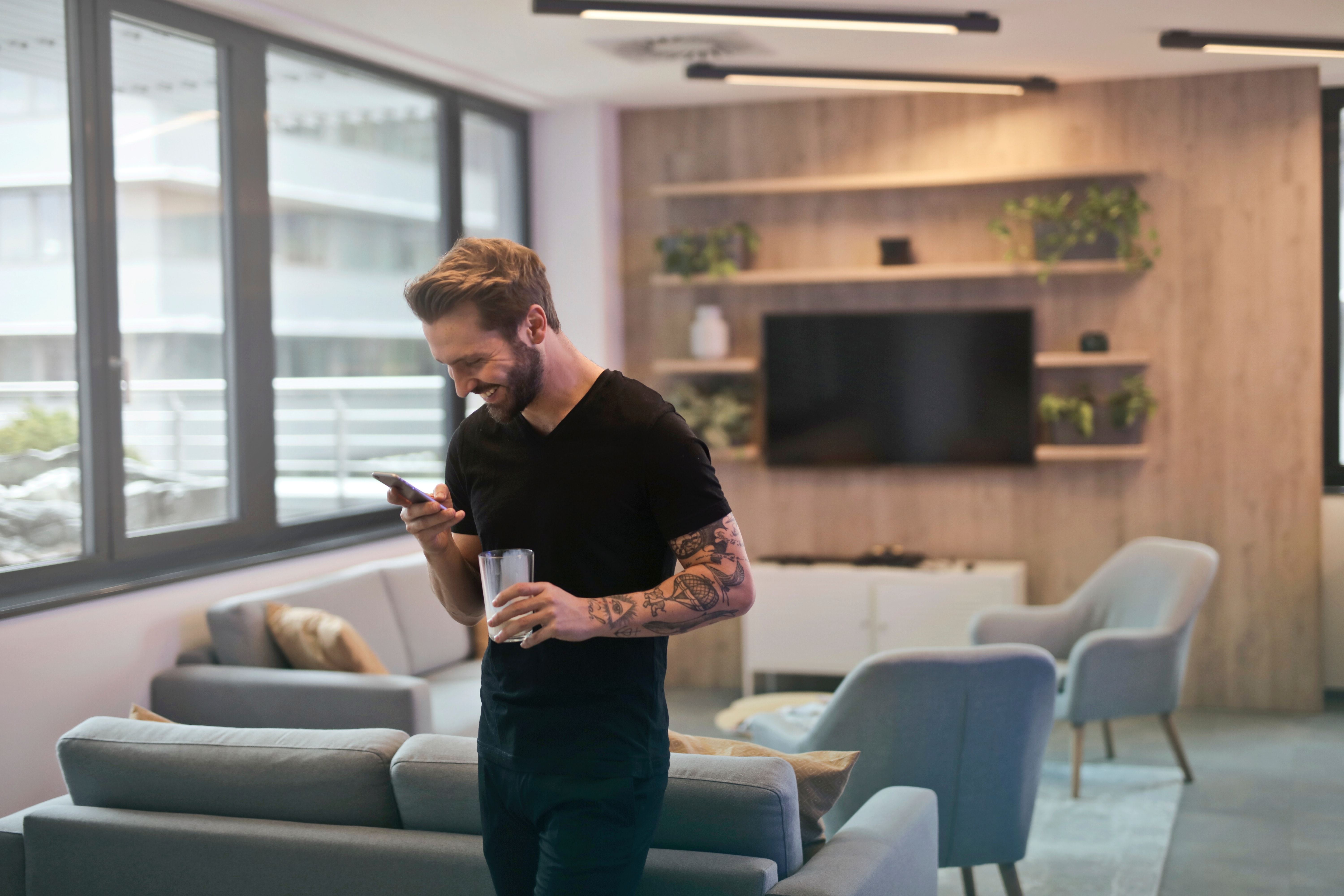 Een man staat in een loungeruimte, kijkt op zijn telefoon en heeft een glas water in zijn hand.