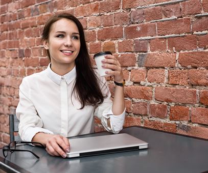 Een vrouw zit op een terras aan een tafel tegen een bakstenen muur aan. De vrouw heeft koffie in haar hand.