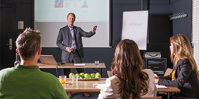 Een man geeft een presentatie aan drie mensen. De drie mensen maken aantekeningen, op tafel liggen appels en staat drinken.