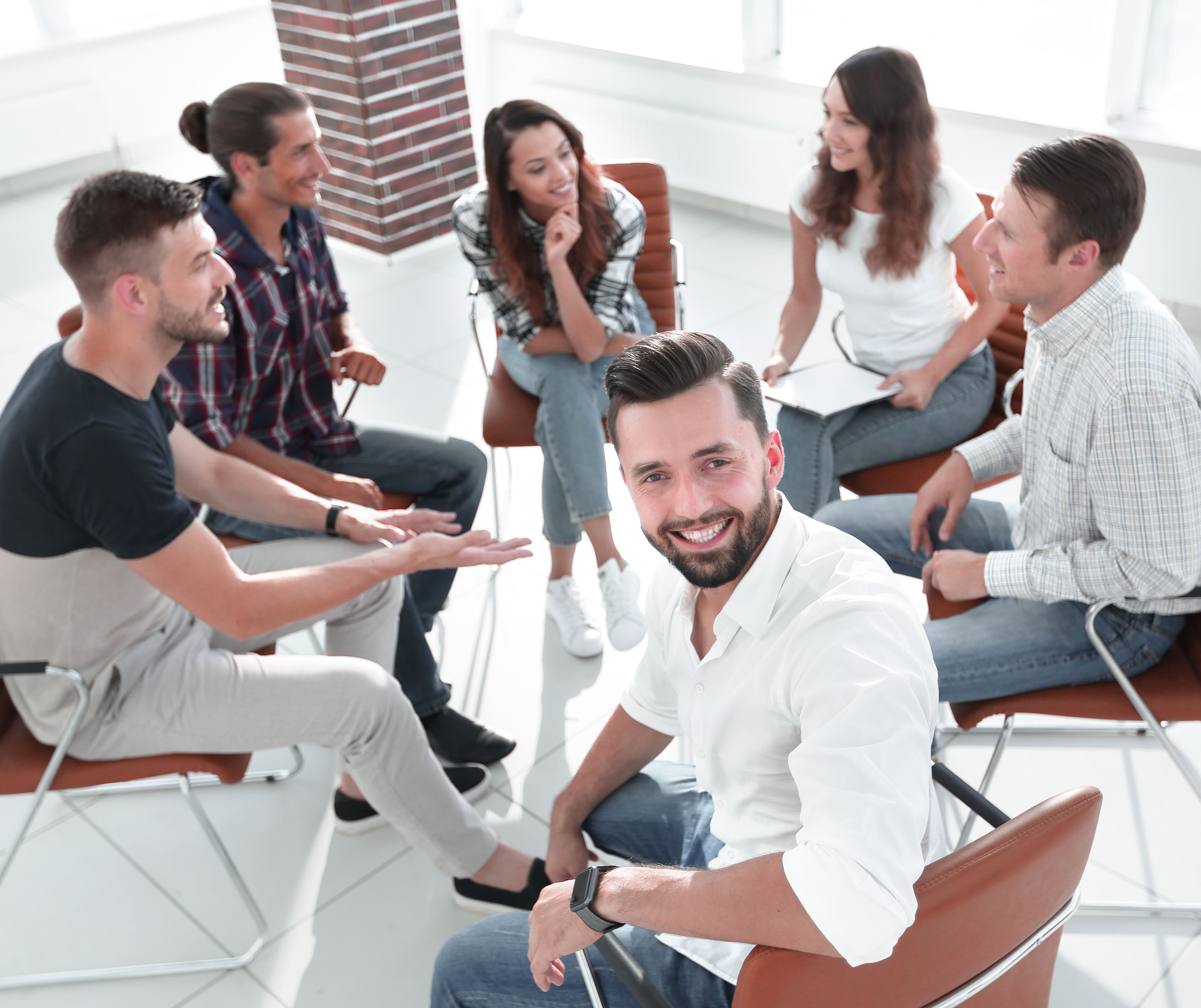 Een groep mensen zitten in een cirkel op bruine stoelen, een man kijkt achterom in de camera. De rest praat met elkaar.
