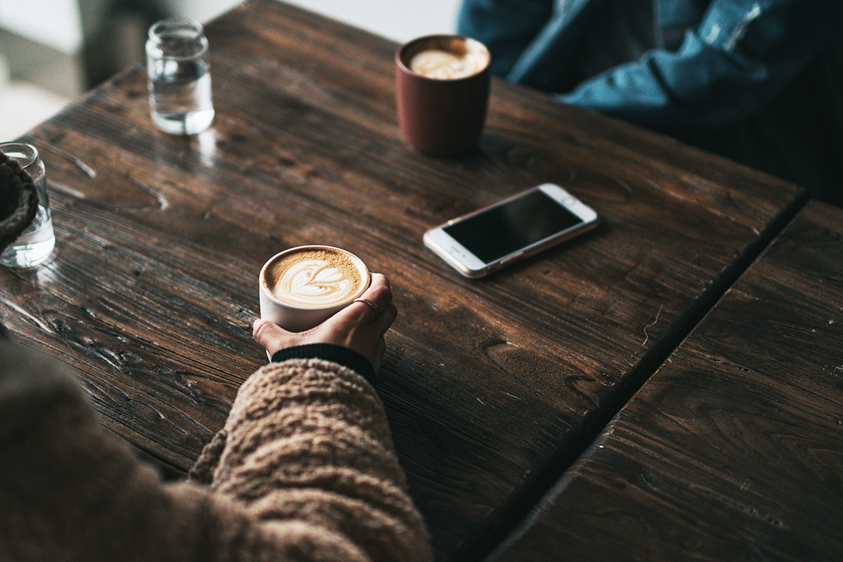 Twee mensen zitten in een cafe koffie te drinken, op tafel staan kopjes koffie en liggen telefoons.