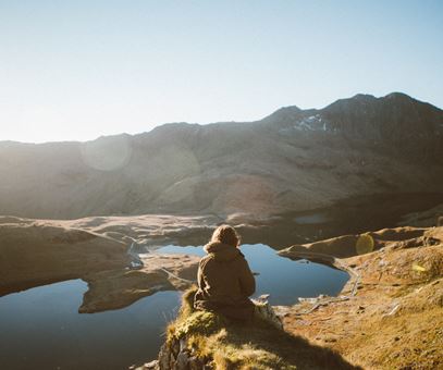 Een vrouw zit op een berg en kijkt uit over een meer en een andere berg.