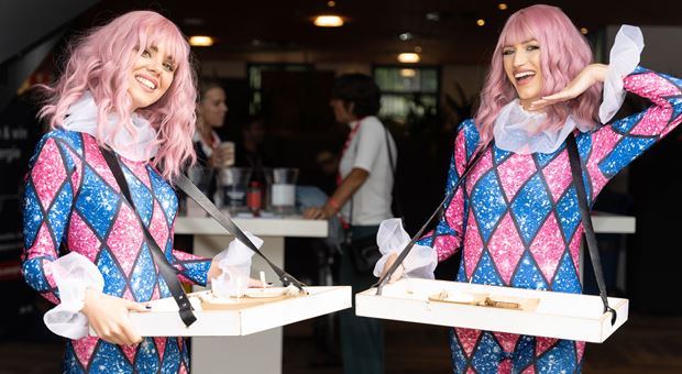 Twee dames in roze met blauw pakken die met een dienblad broodjes rondbrengen. De dames hebben ook een roze pruik op.