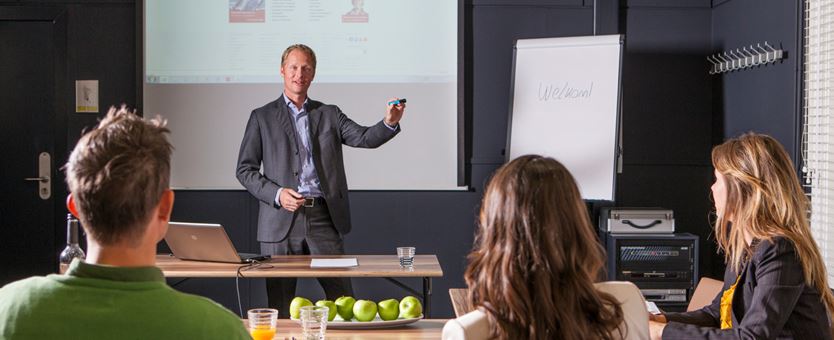 Een man geeft een presentatie aan drie mensen. De drie mensen maken aantekeningen, op tafel liggen appels en staat drinken.