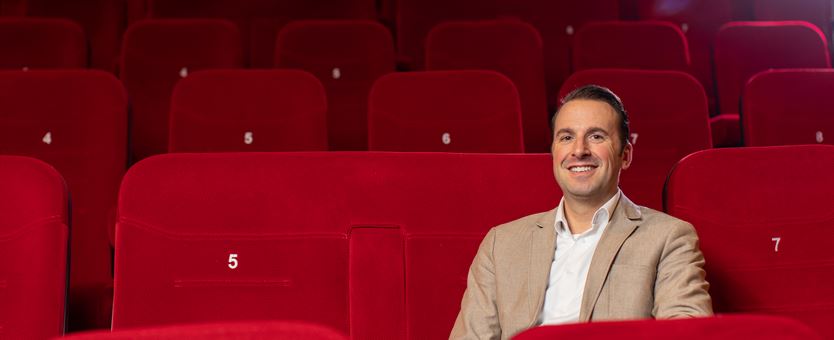 Een man met een witte blouse en beige blazer zit in een lege bioscoopzaal op een bioscoopstoel.