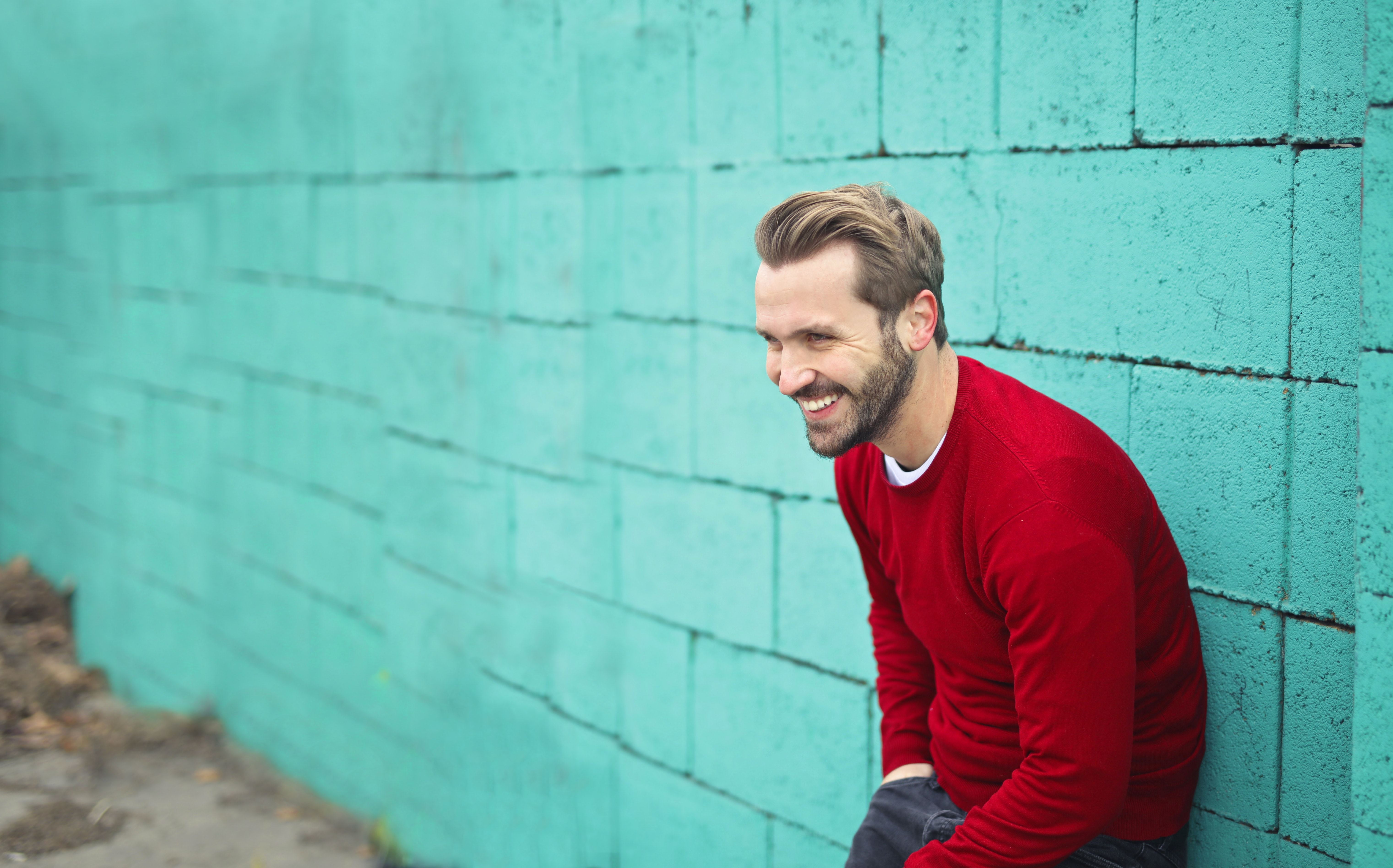 Een man met een rode trui staat tegen een turquoise muur en lacht.