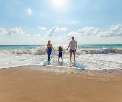 Een moeder, vader en dochter lopen samen hand in hand de zee in. Voor hen komt er veel water omhoog door de golven.