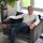 Foto van Robert van Roermund. Een man met grijze haren en een witte blouse zit op een stoel.