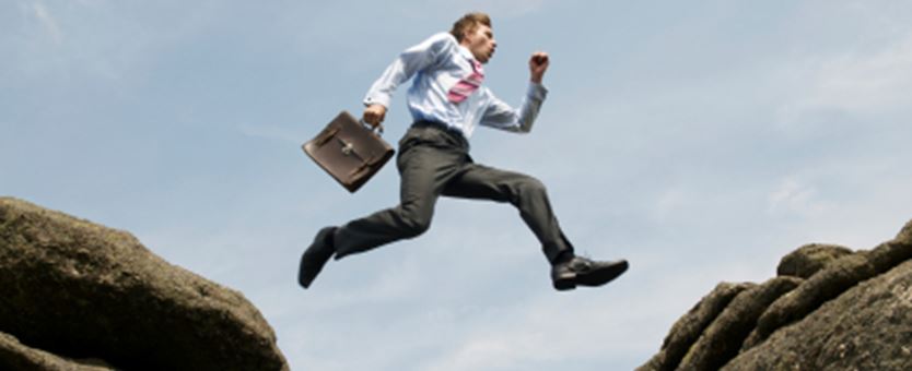 Een man met een blauwe blouse, rode stropdas en laptoptas springt van de ene rots naar de andere.