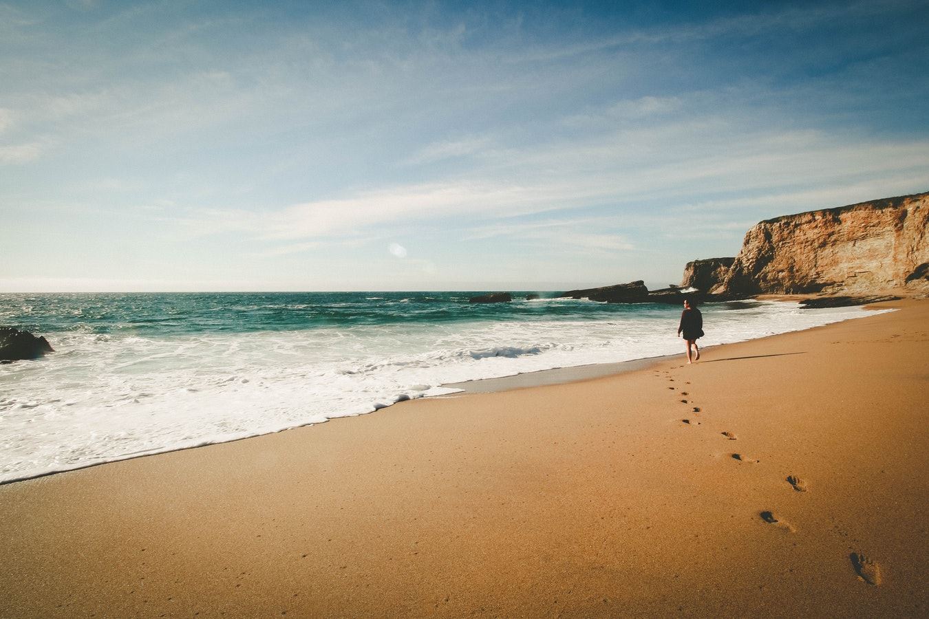 Een vrouw loopt over het strand richting de zee, In het zand staan haar voetafdrukken. Op de achtergrond ligt een klif.