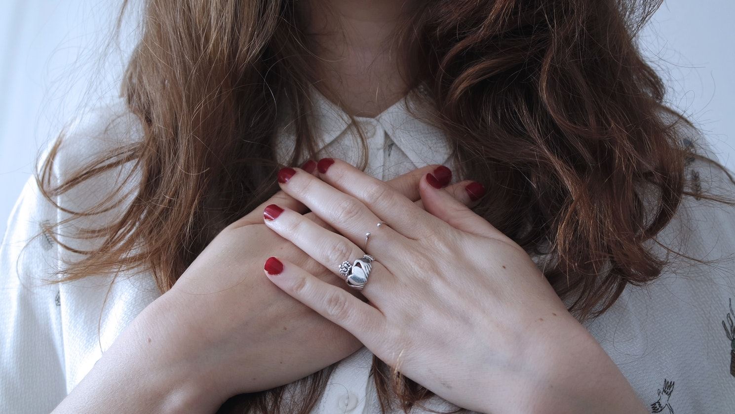 Een vrouw met allebei haar handen op haar hart, ze heeft een witte blouse, zilveren ringen en rode nagellak