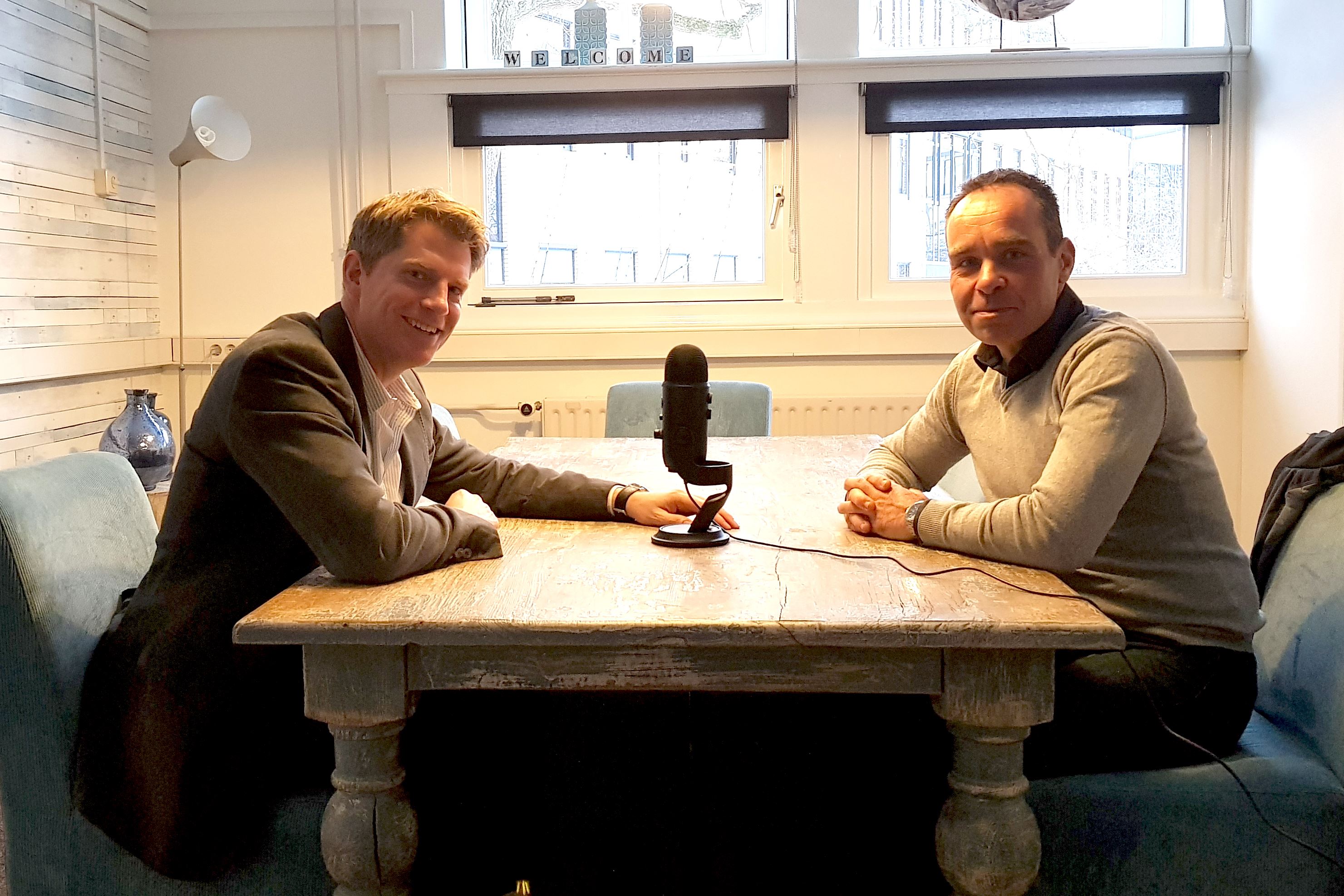 Twee mannen zitten rond een tafel, op de tafel staat een microfoon, ze nemen een podcast op.